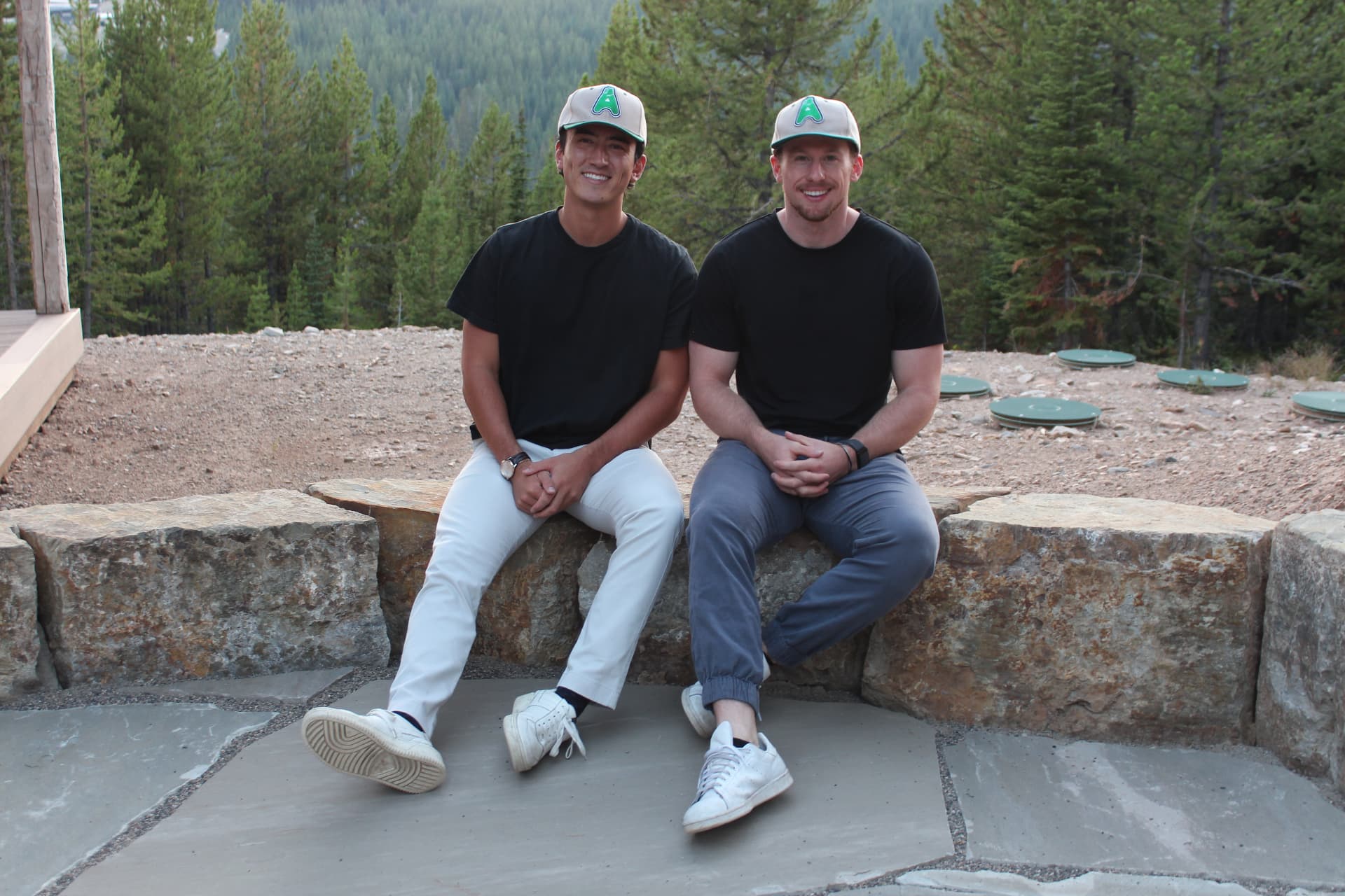 AthletesTogether founders John and Lucas sitting together outdoors in matching black shirts and green logo caps
